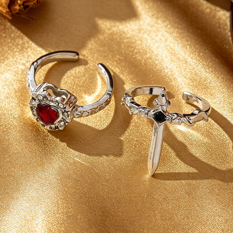 One silver-tone red heart open ring beside one black-accent sword open ring on gold fabric