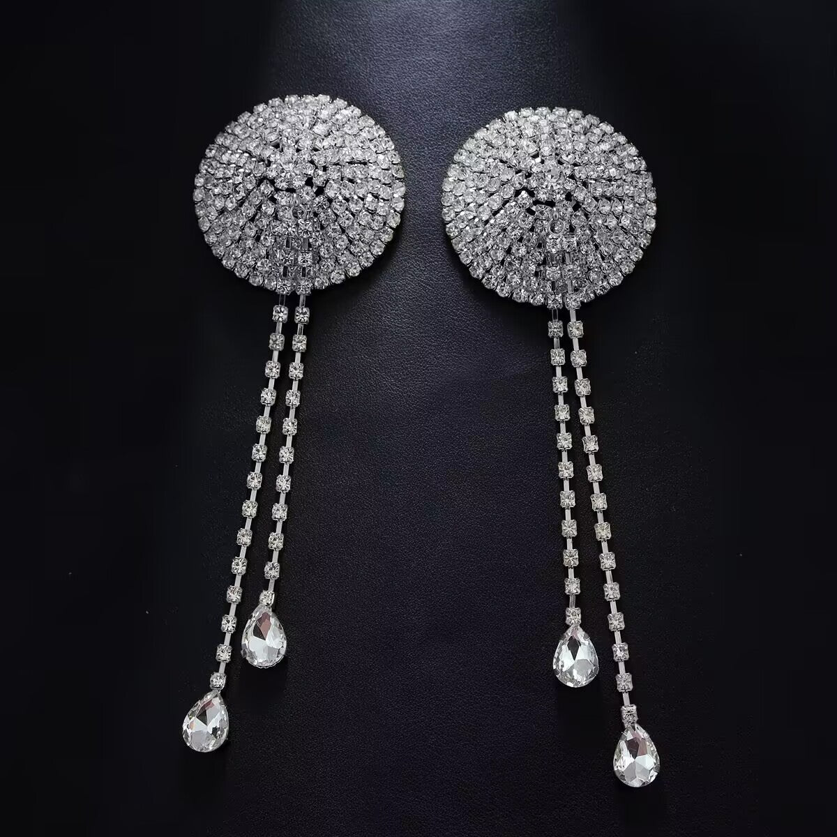 Pair of silver crystal rhinestone tassel nipple covers with teardrop pendants on dark textured background.
