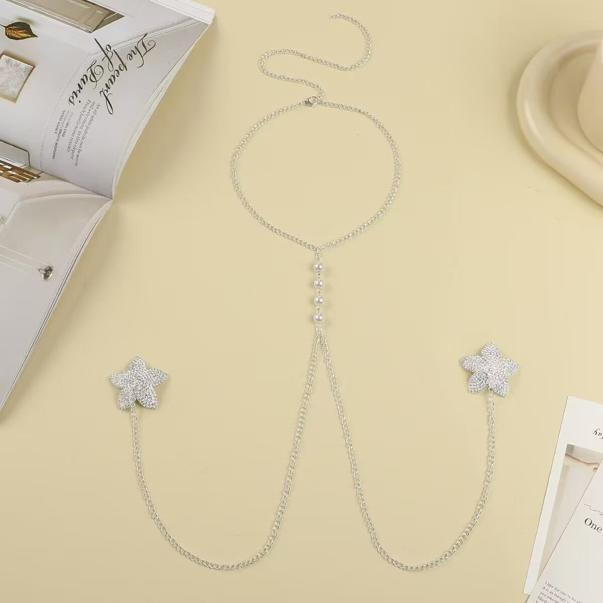 Flatlay of a silver non pierced nipple shield halter neck chain featuring sparkly rhinestone five-point stars and dangling metal chains with inline white pearls.