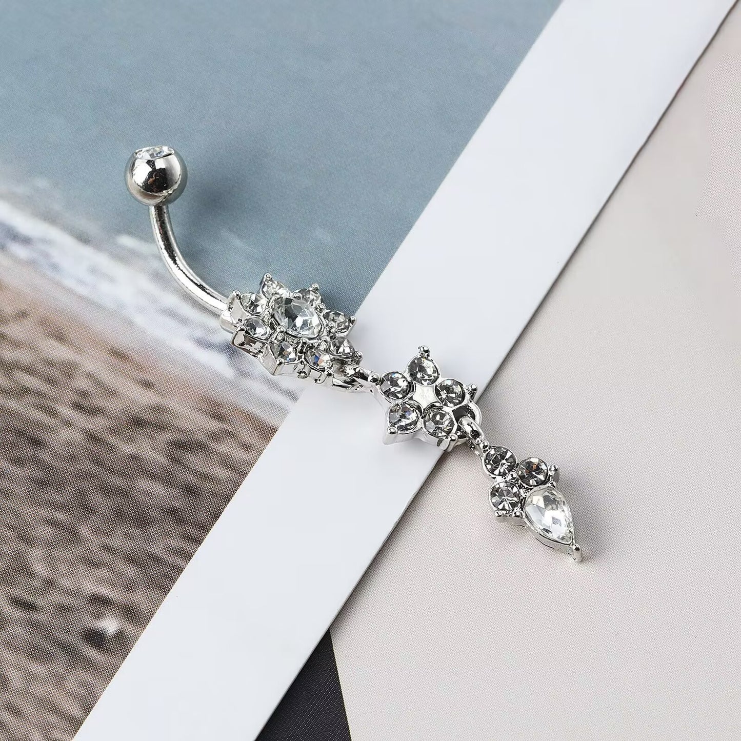Silver starburst crystal dangle belly button piercing jewelry laying on magazine page background.