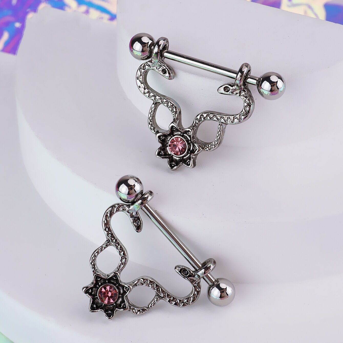 Close-up display of two snake nipple shields with pink gems on a white textured platform, highlighting the intricate metallic details and alternative aesthetic.