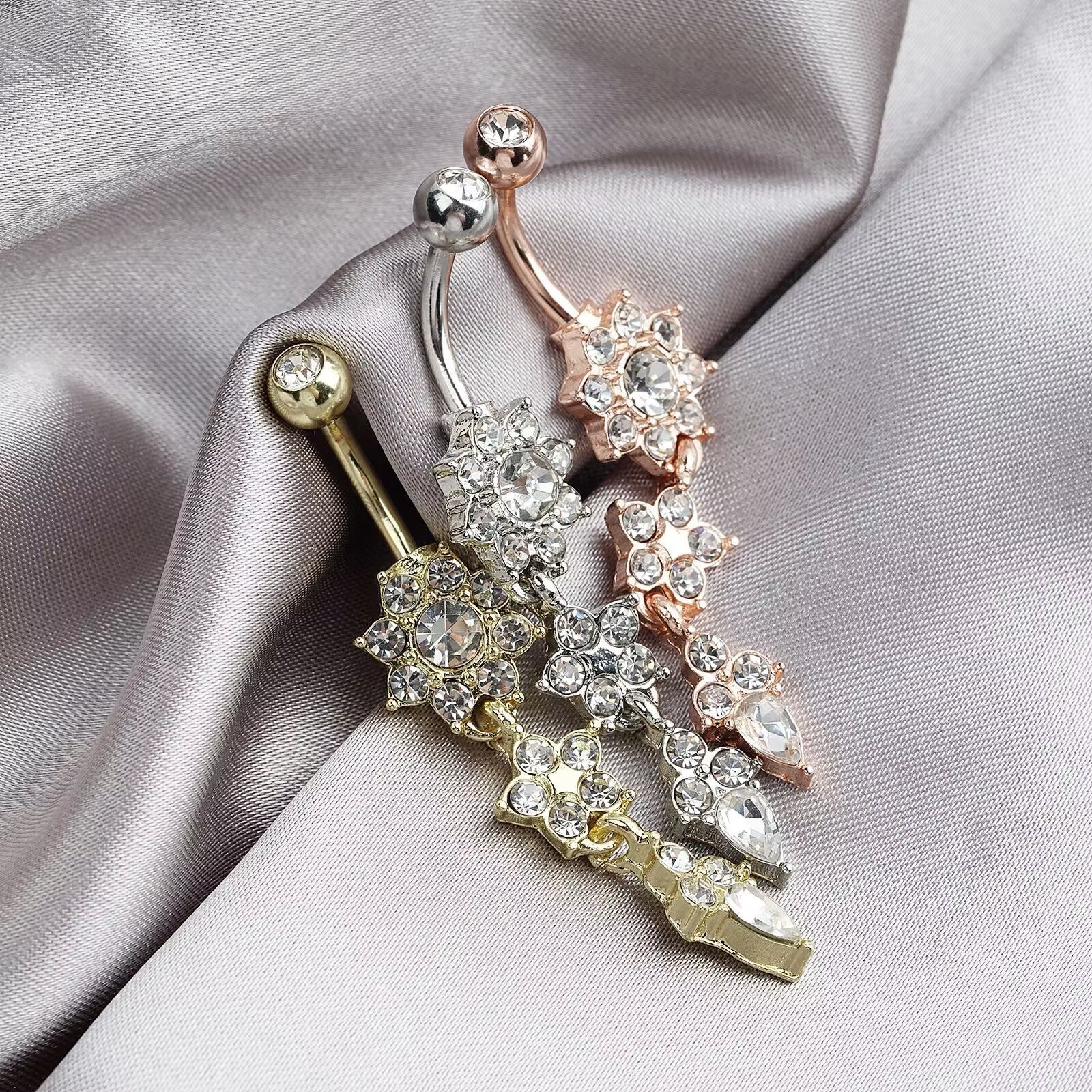 Close-up of crystal flower and teardrop zircon dangle navel piercing rings on soft grey satin background.