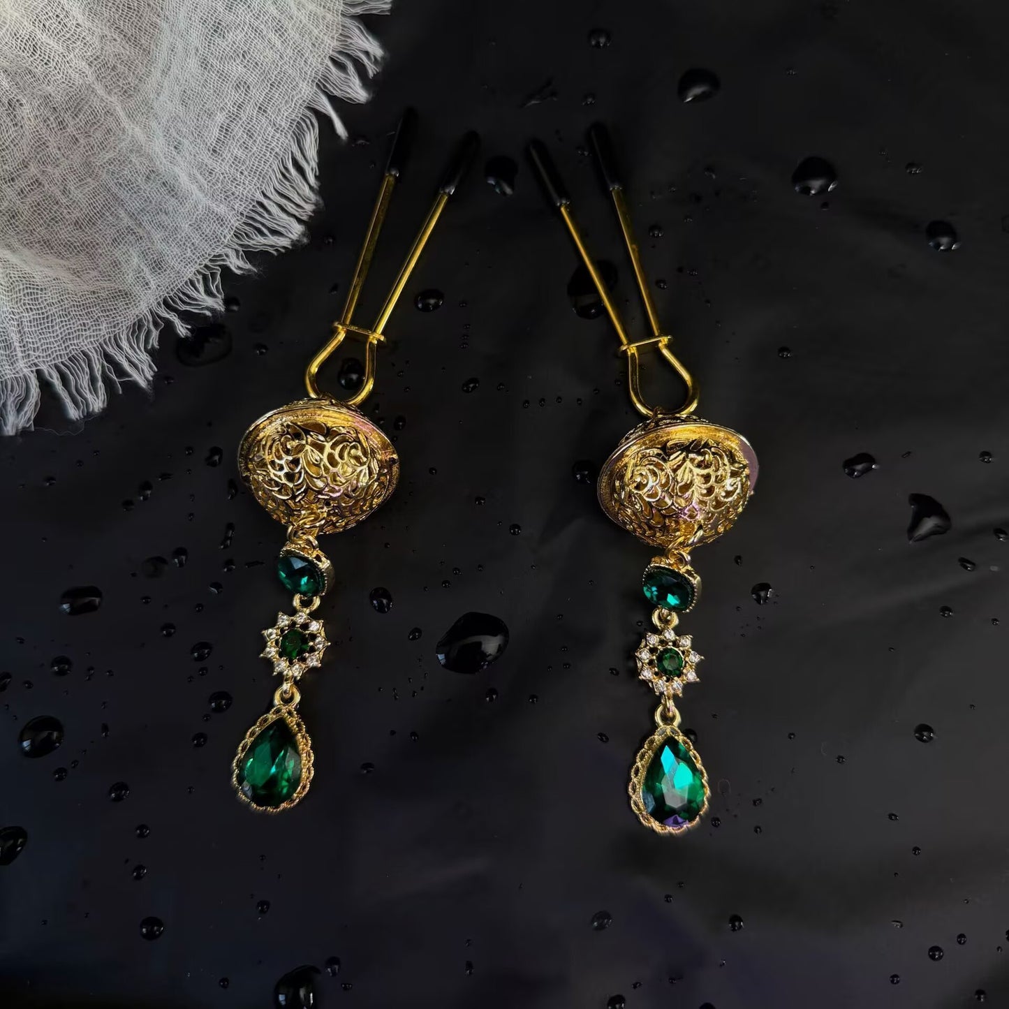 Pair of gold U-shaped long pin nipple clips with protective end caps and emerald green crystal drop accessories.
