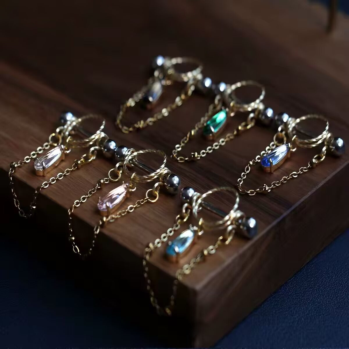 Multi-color waterdrop CZ nipple shield barbells with delicate dangle chains arranged on a dark wooden display.